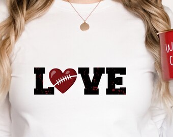 May include: White long-sleeve shirt with the word "LOVE" in black block letters. The "O" is replaced with a football-shaped heart in red and white. A red mug with "warm cozy" written on it.