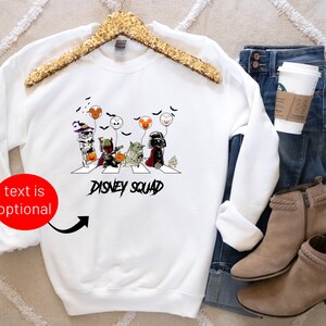 Puede incluir: Sudadera blanca con un gr&aacute;fico de Halloween de personajes de Disney cruzando una calle, con el texto "DISNEY SQUAD". La sudadera se muestra con vaqueros azules, una taza de caf&eacute; y botines color canela.