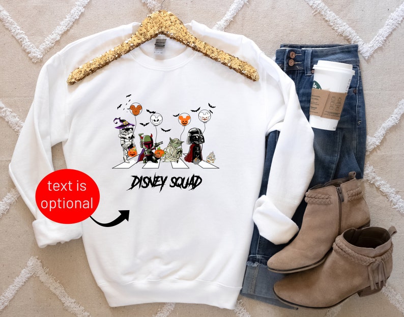 Puede incluir: Sudadera blanca con un gr&aacute;fico de Halloween de personajes de Disney cruzando una calle, con el texto "DISNEY SQUAD". La sudadera se muestra con vaqueros azules, una taza de caf&eacute; y botines color canela.