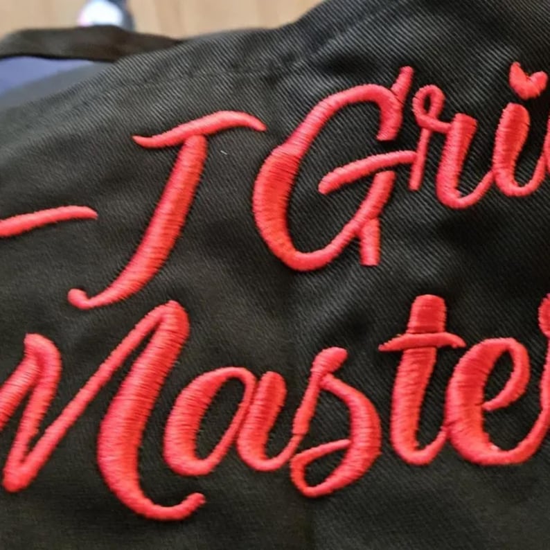 Puede incluir: Primer plano de un delantal negro con las palabras "I Grill Master" bordadas en rojo. El delantal est&aacute; hecho de una tela duradera, perfecto para los entusiastas de la parrilla.
