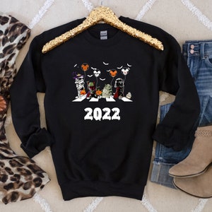 Puede incluir: Sudadera negra de cuello redondo con un gr&aacute;fico de Halloween que representa personajes cruzando una calle, con el a&ntilde;o "2022" en blanco. El dise&ntilde;o incluye murci&eacute;lagos y globos. La sudadera se muestra con vaqueros, botas y una bufanda.