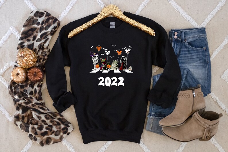 Puede incluir: Sudadera negra de cuello redondo con un gr&aacute;fico de Halloween que representa personajes cruzando una calle, con el a&ntilde;o "2022" en blanco. El dise&ntilde;o incluye murci&eacute;lagos y globos. La sudadera se muestra con vaqueros, botas y una bufanda.