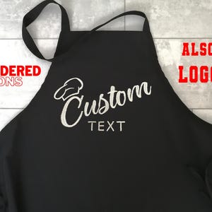 Puede incluir: Delantal negro con texto bordado blanco "Custom TEXT" y un dise&ntilde;o de gorro de chef. El delantal tiene una correa para el cuello y lazos. Las palabras "EMBROIDERED APRONS" y "ALSO LOGO" est&aacute;n en texto rojo.