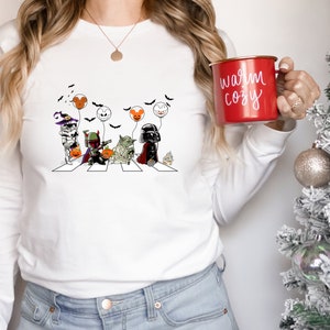 Puede incluir: Camiseta blanca de manga larga con un dise&ntilde;o de parodia de Star Wars con tem&aacute;tica de Halloween. El gr&aacute;fico representa personajes cruzando una calle, sosteniendo globos. Una taza roja con el texto "warm cozy" se sostiene en la mano.