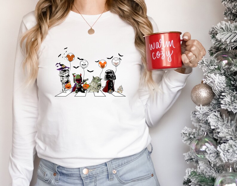 Puede incluir: Camiseta blanca de manga larga con un dise&ntilde;o de parodia de Star Wars con tem&aacute;tica de Halloween. El gr&aacute;fico representa personajes cruzando una calle, sosteniendo globos. Una taza roja con el texto "warm cozy" se sostiene en la mano.