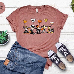 May include: Heather mauve t-shirt with a Halloween design of cartoon skeletons holding character-shaped balloons. The shirt is styled with blue jeans and black trainers. The text "Heather Mauve" is in the top left corner.