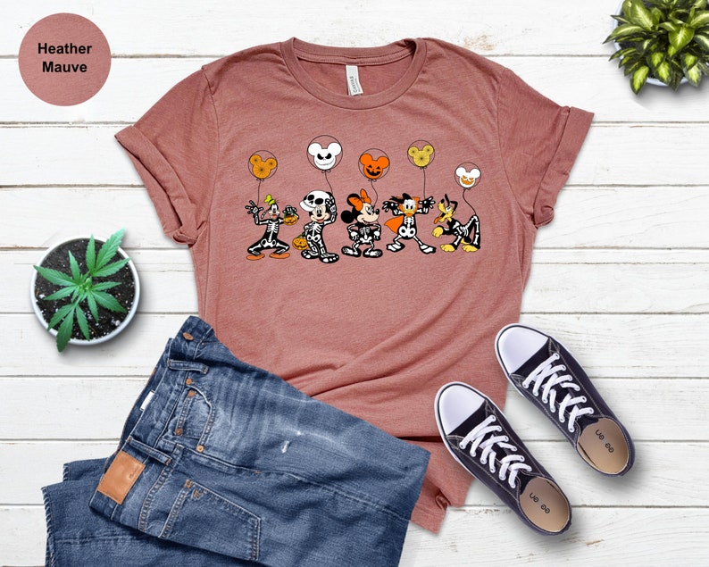 May include: Heather mauve t-shirt with a Halloween design of cartoon skeletons holding character-shaped balloons. The shirt is styled with blue jeans and black trainers. The text "Heather Mauve" is in the top left corner.