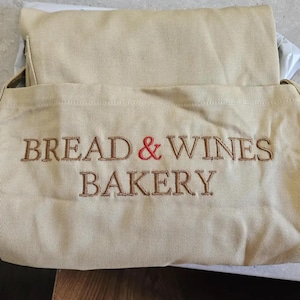 Puede incluir: Bolso de mano de lona beige con el texto bordado "BREAD & WINES BAKERY" en hilo marr&oacute;n, con una & roja. La bolsa est&aacute; doblada.