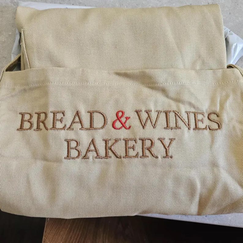Puede incluir: Bolso de mano de lona beige con el texto bordado "BREAD & WINES BAKERY" en hilo marr&oacute;n, con una & roja. La bolsa est&aacute; doblada.