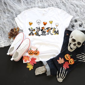May include: White t-shirt with a Halloween cartoon graphic of characters holding balloons. The image includes white trainers, a pair of dark blue jeans, a skull, and decorative pumpkins and leaves, arranged on a black surface.