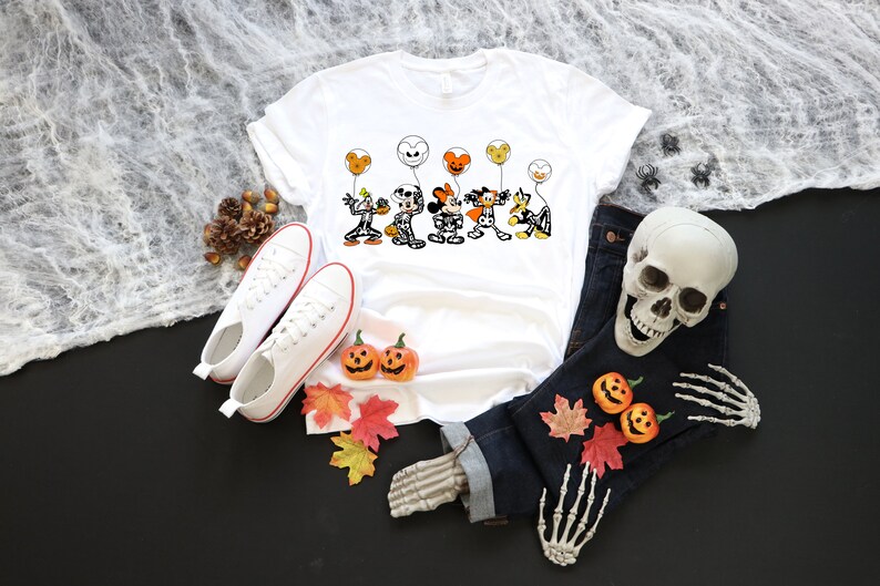 May include: White t-shirt with a Halloween cartoon graphic of characters holding balloons. The image includes white trainers, a pair of dark blue jeans, a skull, and decorative pumpkins and leaves, arranged on a black surface.