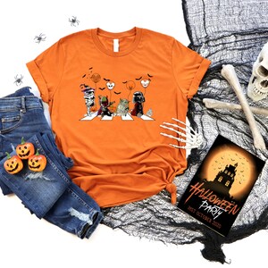 Puede incluir: Camiseta naranja con un gr&aacute;fico de Halloween que muestra personajes cruzando una calle, con calabazas y vaqueros. Tambi&eacute;n hay una invitaci&oacute;n a una fiesta de Halloween y un esqueleto decorativo.