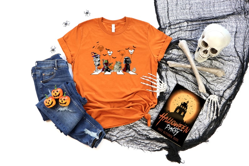 Puede incluir: Camiseta naranja con un gr&aacute;fico de Halloween que muestra personajes cruzando una calle, con calabazas y vaqueros. Tambi&eacute;n hay una invitaci&oacute;n a una fiesta de Halloween y un esqueleto decorativo.
