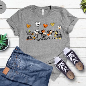 May include: A heather light grey t-shirt with a Halloween design featuring Disney characters as skeletons, holding balloon-shaped heads. The characters include Goofy, Mickey Mouse, Minnie Mouse, and Donald Duck. The shirt is styled with blue jeans and black trainers.
