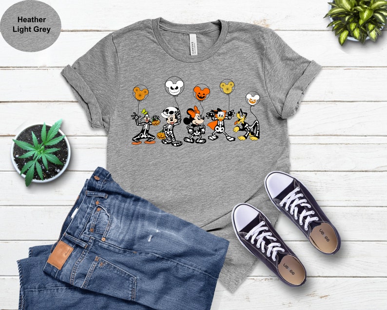 May include: A heather light grey t-shirt with a Halloween design featuring Disney characters as skeletons, holding balloon-shaped heads. The characters include Goofy, Mickey Mouse, Minnie Mouse, and Donald Duck. The shirt is styled with blue jeans and black trainers.