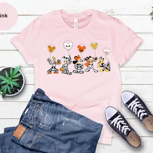 May include: Pale pink t-shirt with a Halloween cartoon graphic of characters in skeleton costumes holding balloons. The word "Pink" is in a pink circle. The image also includes blue jeans and black trainers.