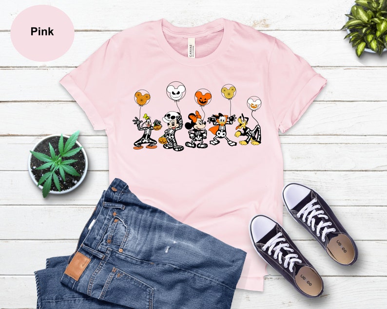 May include: Pale pink t-shirt with a Halloween cartoon graphic of characters in skeleton costumes holding balloons. The word "Pink" is in a pink circle. The image also includes blue jeans and black trainers.