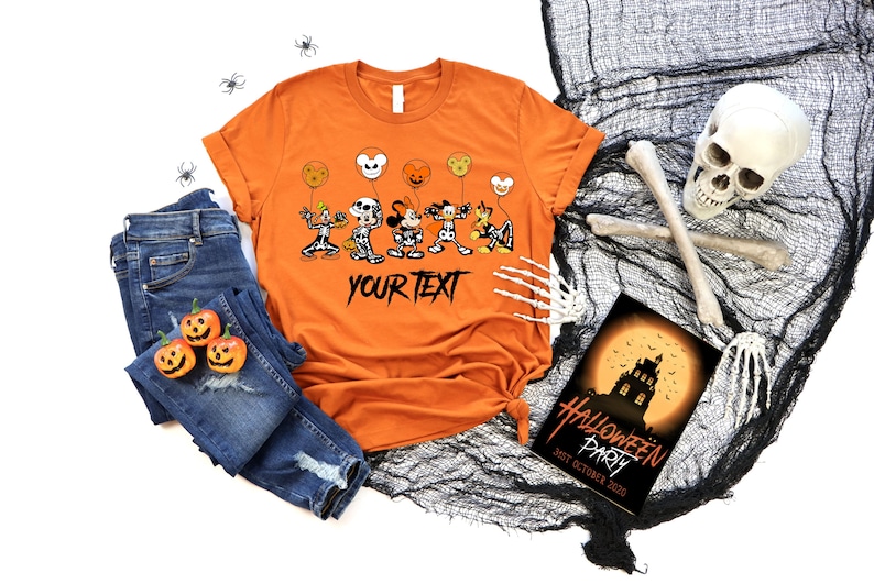 May include: Orange t-shirt with a Halloween cartoon design of characters holding balloons and the text "YOUR TEXT". The image includes blue jeans, decorative pumpkins, and a Halloween party invitation. The scene is Halloween themed.