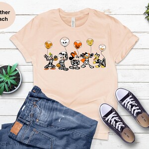 May include: A peach-coloured t-shirt with a Halloween cartoon design. The graphic depicts cartoon characters in costumes, holding balloons. The shirt is styled with blue jeans and black trainers.
