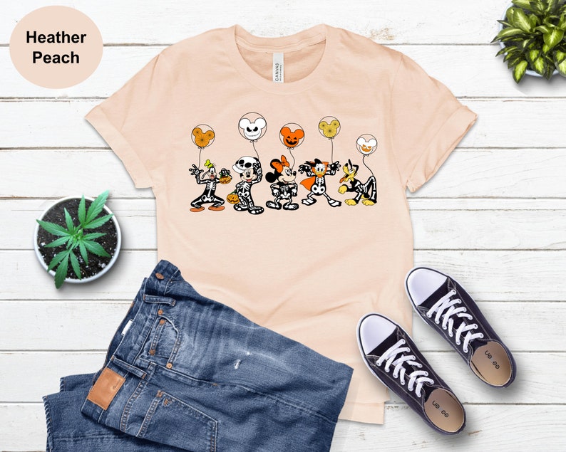 May include: A peach-coloured t-shirt with a Halloween cartoon design. The graphic depicts cartoon characters in costumes, holding balloons. The shirt is styled with blue jeans and black trainers.