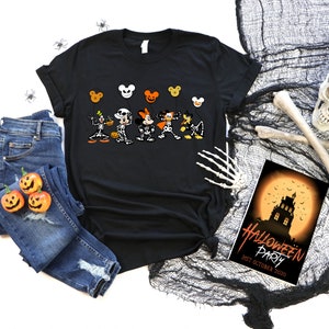 May include: Black t-shirt with a Halloween cartoon character design, paired with blue jeans and pumpkin decorations. A Halloween party invitation and spooky decorations are also visible. The scene evokes a festive atmosphere.