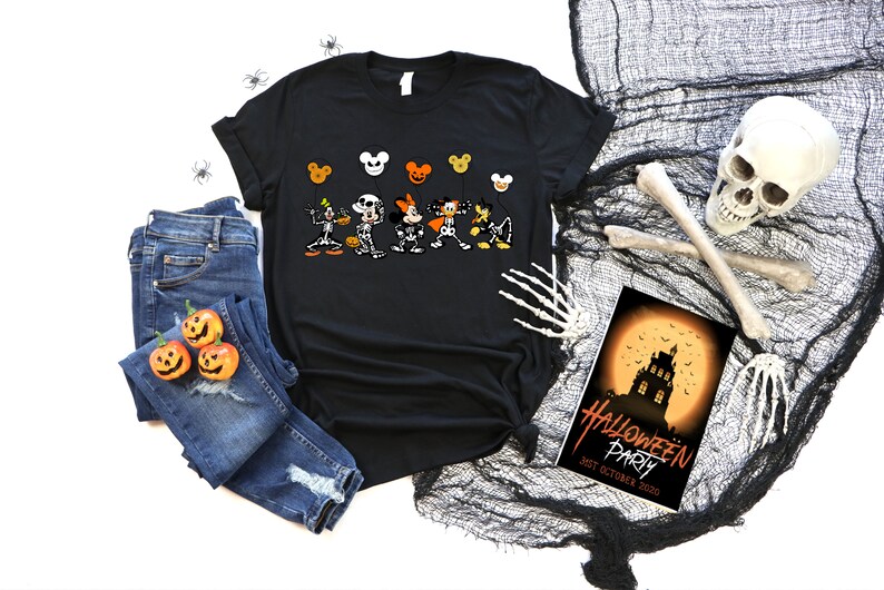 May include: Black t-shirt with a Halloween cartoon character design, paired with blue jeans and pumpkin decorations. A Halloween party invitation and spooky decorations are also visible. The scene evokes a festive atmosphere.