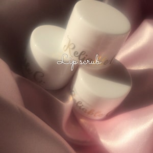 Puede incluir: Tres frascos blancos de exfoliante labial con letras doradas que dicen "Reliable Lip Scrub". Los frascos están sobre una tela de satén rosa.