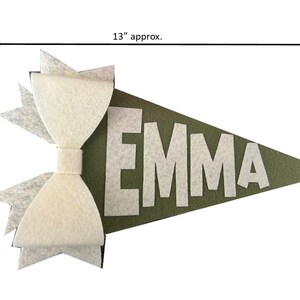 Może przedstawiać: Zielony proporzec z filcu z białą kokardą z filcu i imieniem "EMMA" w białych filcowych literach. Proporzec ma około 33 cm długości i 18 cm wysokości.