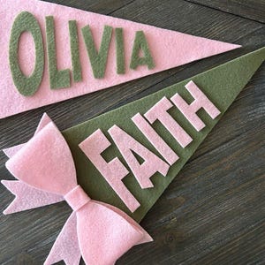 Może przedstawiać: Dwa flagi z filcu z imionami Olivia i Faith w zielonych literach na r&oacute;żowym i zielonym tle. R&oacute;żowa kokardka z filcu znajduje się na pierwszym planie.