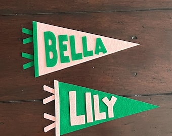 May include: Two triangular felt pennants with ties on the left side. The top pennant is light pink with the name BELLA in green letters. The bottom pennant is green with the name LILY in light pink letters. Both pennants feature contrasting colors.