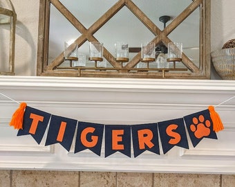 May include: A black felt banner with orange felt letters spelling "TIGERS" and a paw print. The banner has orange tassels on each end.