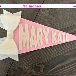 Może przedstawiać: R&oacute;żowa flaga z filcu w kształcie proporca z białą kokardą z filcu i imieniem "MARY KATE" w białych literach z filcu. Flaga ma około 33 cm długości i 18 cm szerokości.