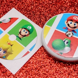 May include: Two round stickers featuring characters from the Super Mario Bros. video game franchise. The stickers are printed with a colorful grid pattern and feature Mario, Luigi, Peach, and Yoshi.