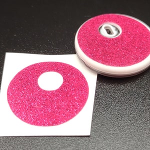 May include: A pink glitter sticker with a white circle in the center. The sticker is on a white backing.