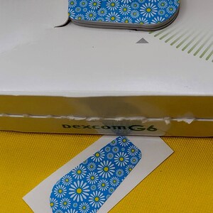 May include: A white sticker with a blue background and a white daisy pattern. The sticker is designed to fit a Dexcom G6 continuous glucose monitor.