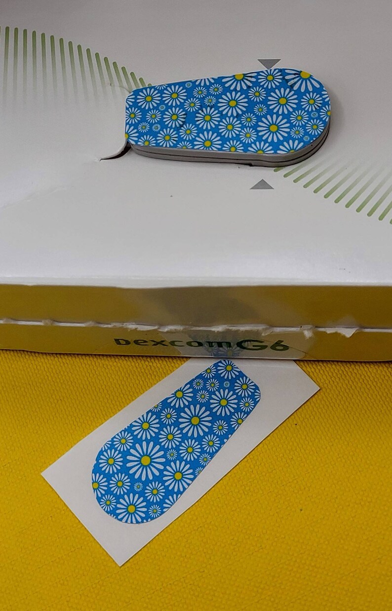 May include: A white sticker with a blue background and a white daisy pattern. The sticker is designed to fit a Dexcom G6 continuous glucose monitor.