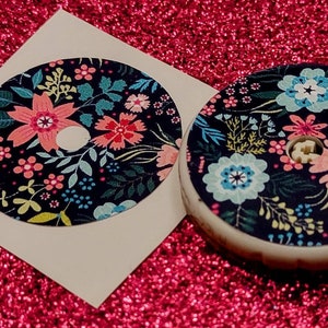 May include: Two round stickers with a floral pattern on a dark blue background. The stickers feature pink, blue, and yellow flowers with green leaves. One sticker is on a white backing, while the other is on a white round base.