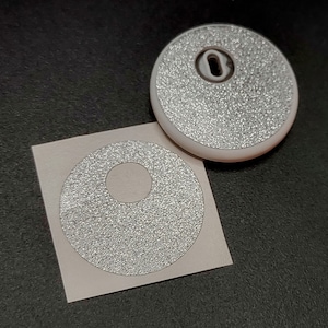 May include: Two white, round, adhesive-backed, reflective stickers with a hole in the center. The stickers have a textured, sparkly surface.