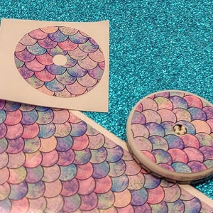 May include: A round, mermaid-scale design in pastel colors of pink, purple, and blue. The design is on a sticker and a small, round object. The background is a sparkly, teal surface.