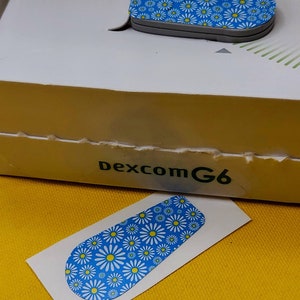 May include: A blue and white patterned sticker with a daisy design. The sticker is for a Dexcom G6 continuous glucose monitor.