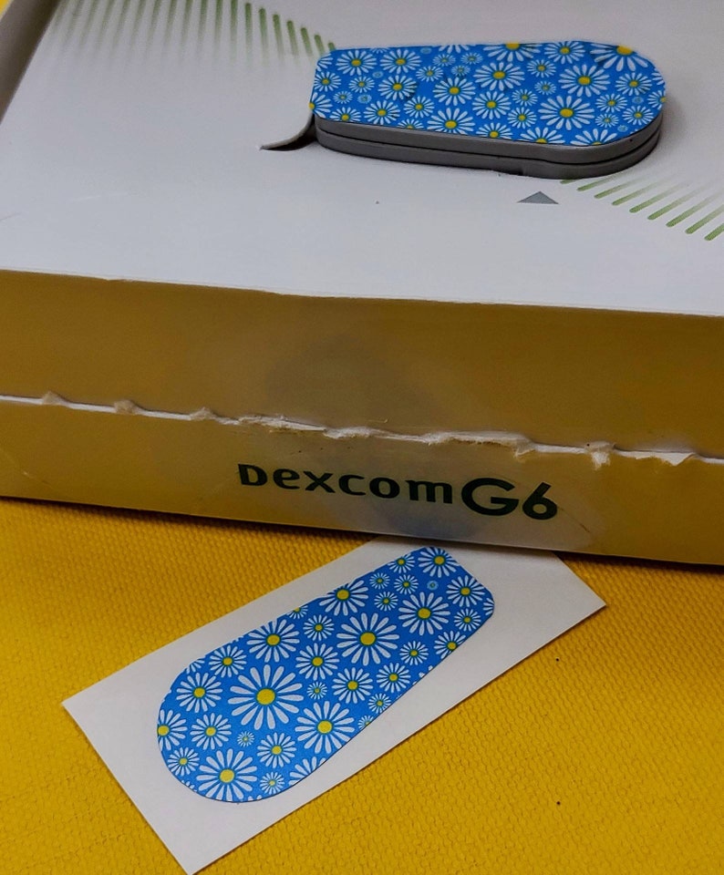 May include: A blue and white patterned sticker with a daisy design. The sticker is for a Dexcom G6 continuous glucose monitor.