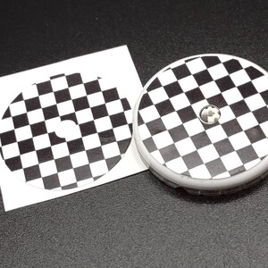 May include: Two black and white checkered round stickers. One sticker is on a white backing sheet and the other is on a round, white, plastic base.