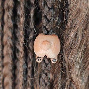 May include: Close-up of brown and black braided hair with a wooden hair bead. The bead is light brown with a carved design. The hair has a mix of textures, including braids and loose curls, creating a natural and textured look.