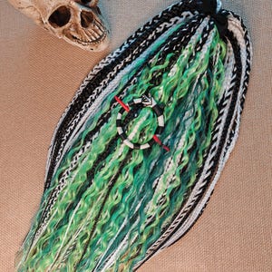 May include: A set of dreadlocks in shades of green, black, and white, with a decorative ring featuring a snake design. The dreadlocks are styled with a mix of straight and wavy textures. A skull is visible in the background.