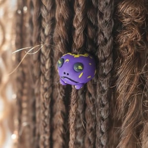 Könnte beinhalten: Nahaufnahme von braunem und blondem geflochtenem Haar mit einem lila Haaraccessoire. Das Accessoire ist ein kleiner Cartoon-Frosch mit gelben Akzenten und einem traurigen Gesichtsausdruck.