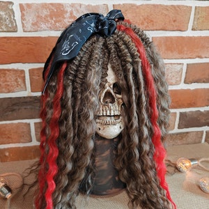 Op de afbeelding: Een schedel met lange bruine dreadlocks met rode strepen, die een zwarte bandana draagt ​​die bovenop het hoofd in een strik is geknoopt.
