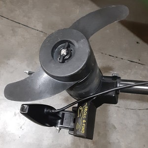 May include: A black trolling motor with two propeller blades. The motor has a black housing and a "TROLL & FIND" label. The motor is designed for use in water and is likely used for fishing or boating.