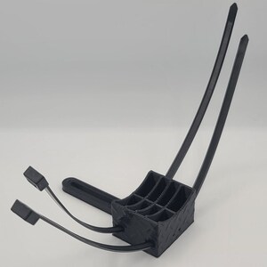 May include: A black 3D-printed object with a square base and a grid-like interior. Two curved black zip ties extend upwards, and two more extend forward. The object is designed to hold multiple items.