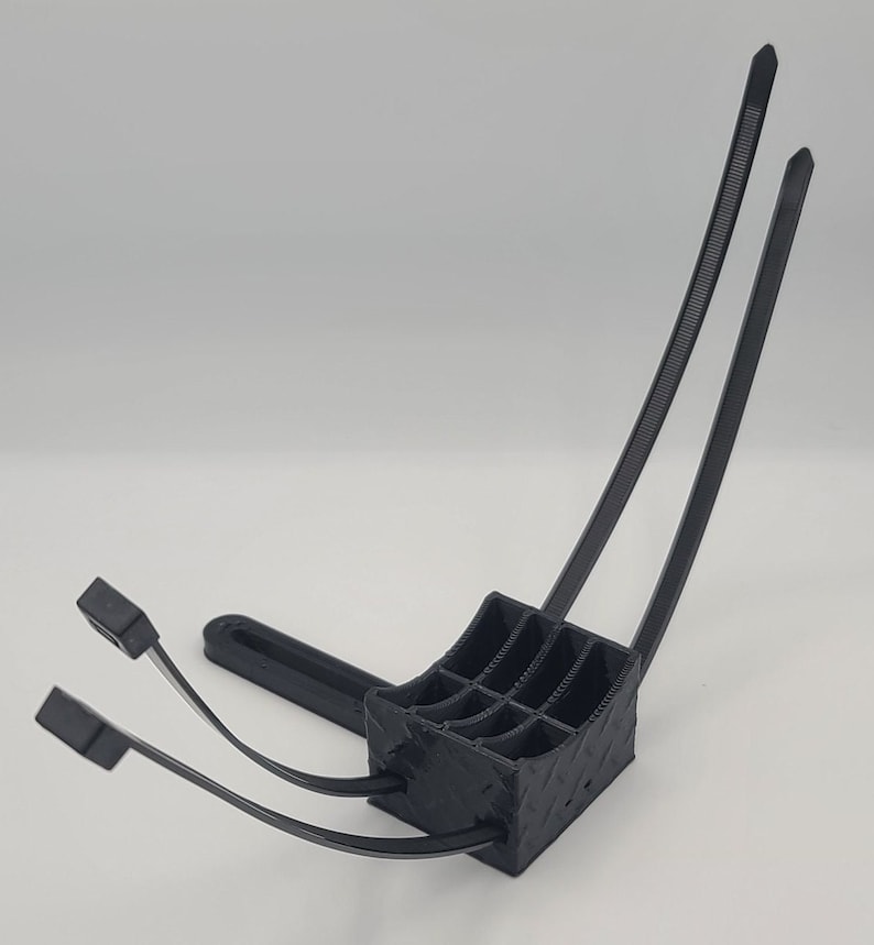 May include: A black 3D-printed object with a square base and a grid-like interior. Two curved black zip ties extend upwards, and two more extend forward. The object is designed to hold multiple items.