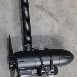 May include: A black trolling motor with a propeller, fin, and a ribbed, flexible shaft. The motor has a streamlined design and is mounted on a bracket. The image is taken against a gray concrete surface.